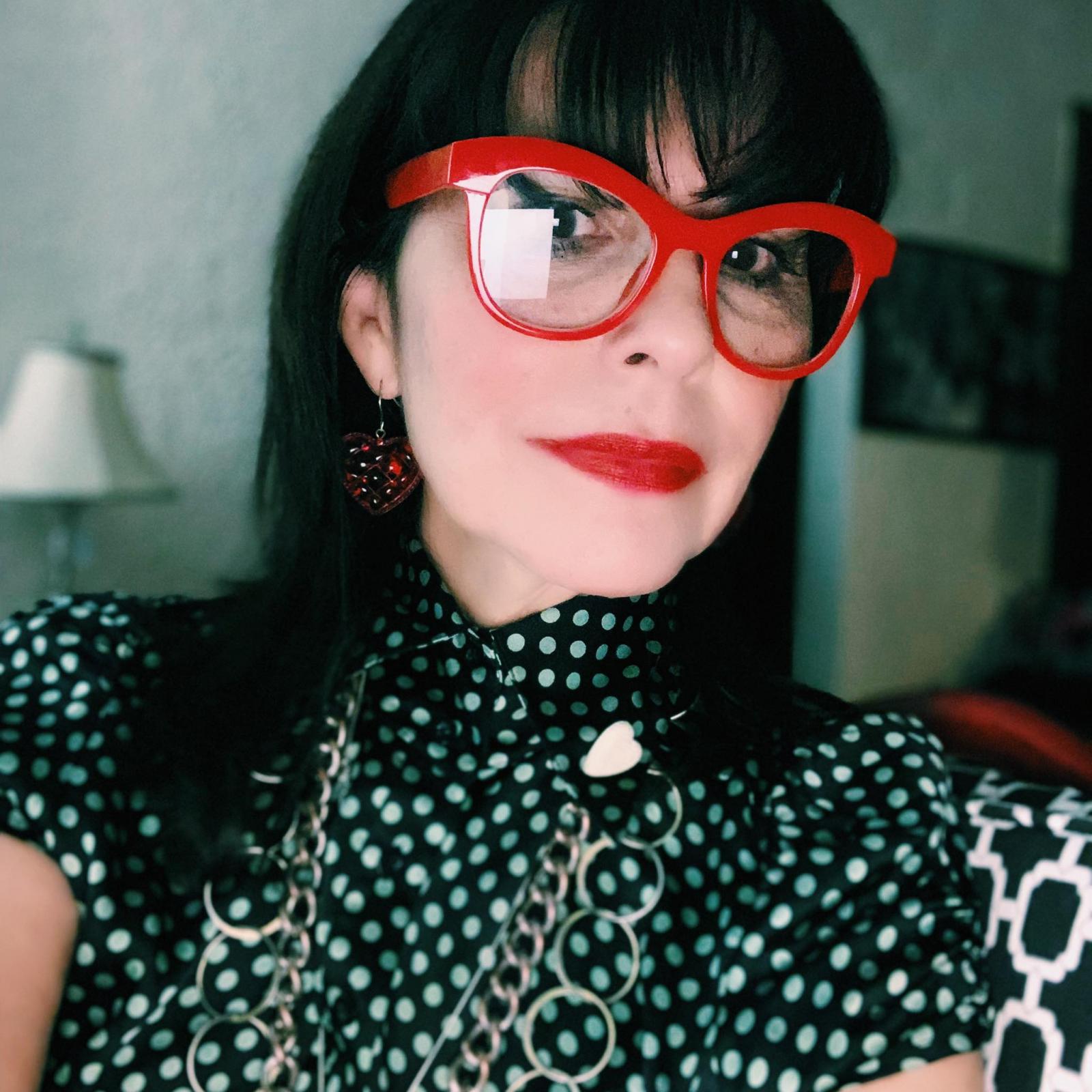Woman with black hair and red glasses in a polka dot shirt with collar smirks at camera