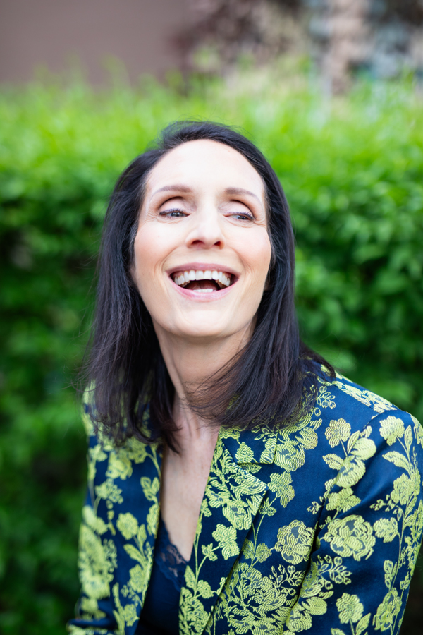 Woman in green shirt in front of green foliage looks into the sky and laughs
