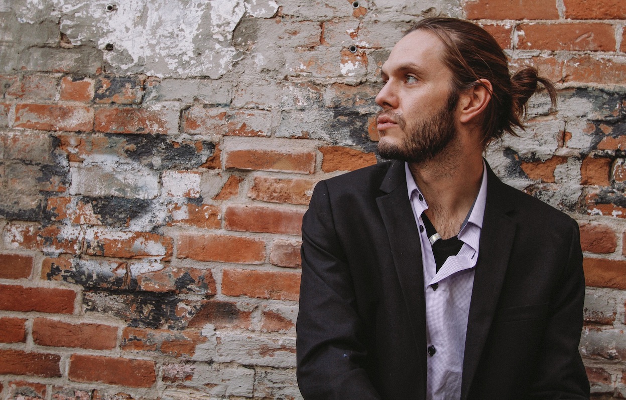 Man in suit jacket and hair in a ponytail faces his right in front of a brick wall