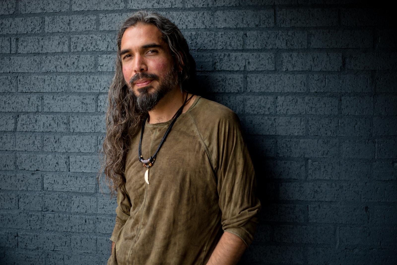 Manwith long dark hair in brown shirt leans against a wall