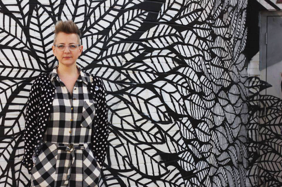 Person in black and white plaid and glasses  stands in front of black and white mural 