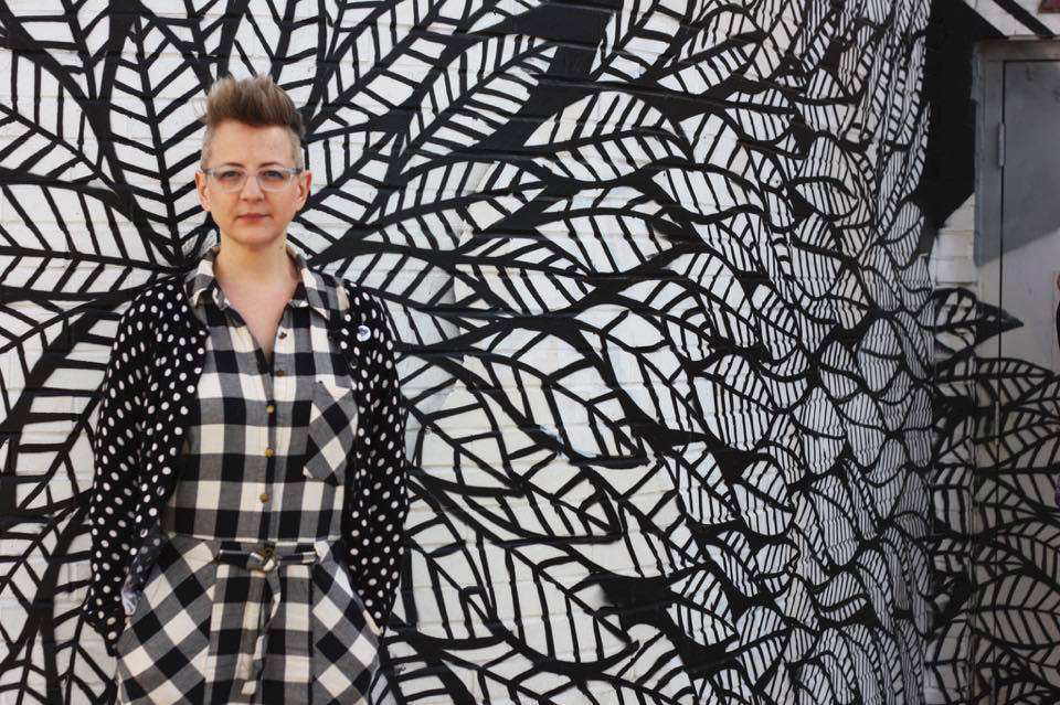 Person in black and white plaid and glasses stands in front of black and white mural