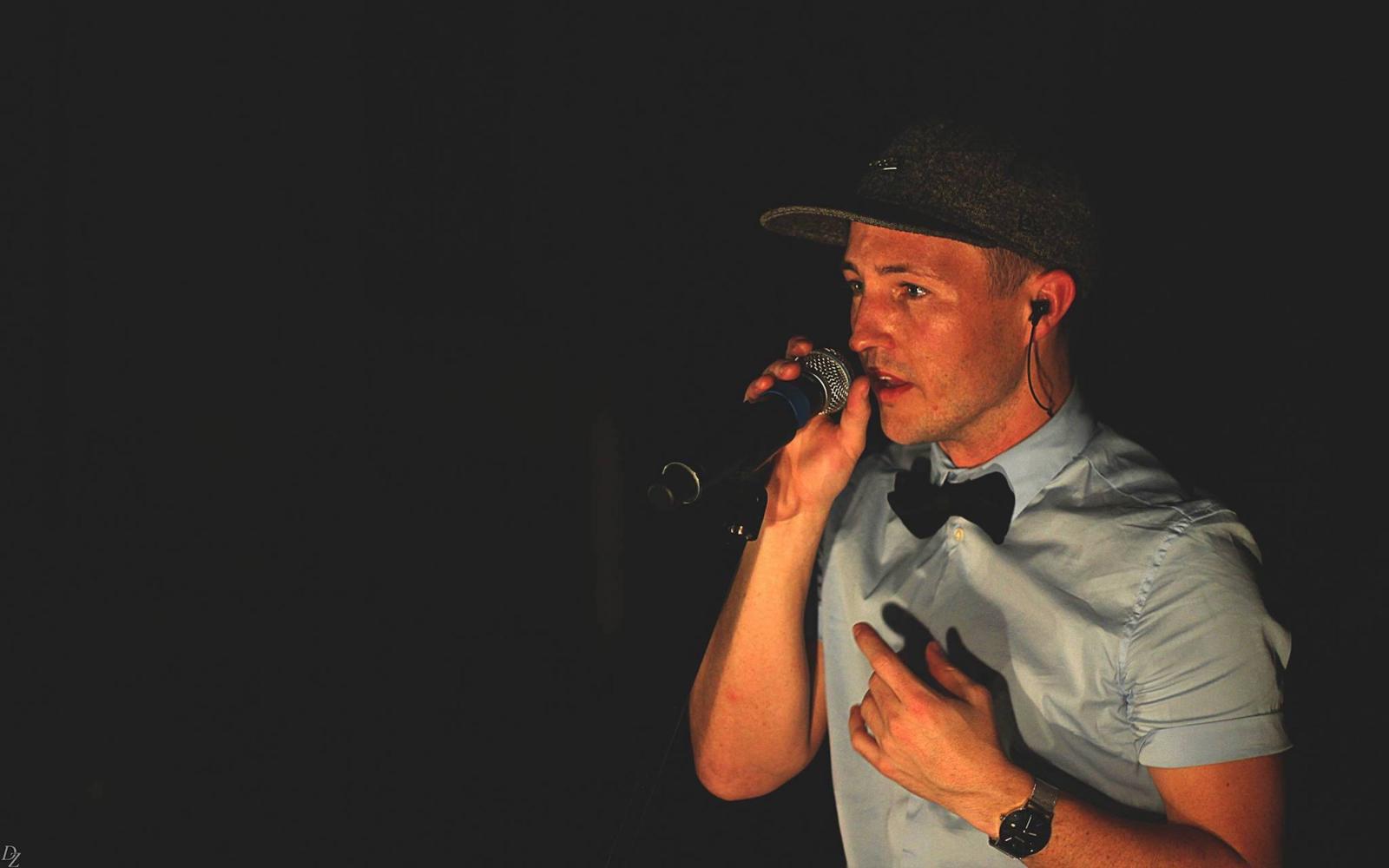 Man in baseball cap beatboxes into microphone. Dark background